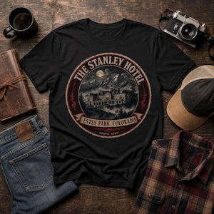 May include: A black short-sleeve t-shirt featuring a circular graphic print of The Stanley Hotel in Estes Park, Colorado. The design includes a detailed illustration of the building set against mountains and a full moon, with text framing the image.