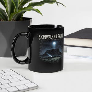 Pode incluir: Uma caneca de cerâmica preta com uma estampa gráfica de uma mesa sob um céu noturno com uma lua brilhante e um feixe de luz. O texto SKINWALKER RANCH e UINTAH BASN, UTAH está escrito em letras brancas acima da ilustração da paisagem.