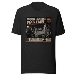 May include: A charcoal gray t-shirt featuring a graphic print with the text MOON LANDING WAS FAKE. The design depicts a film set with a person in a director chair, a camera, and an astronaut, including additional text like IT'S ALL A SET.