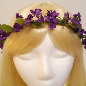 May include: A floral crown featuring vibrant purple flowers and green leaves. The crown is set against a backdrop of long, blonde hair, creating a striking contrast. The flowers are clustered, adding a touch of elegance.