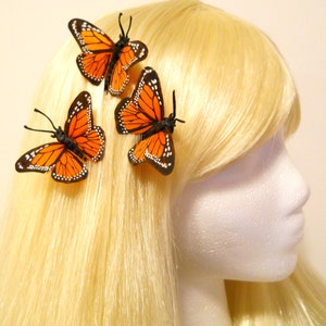 May include: Three orange and black butterfly hair clips are attached to blonde hair. The butterfly wings have a detailed pattern with white spots. The hair clips are a decorative accessory.