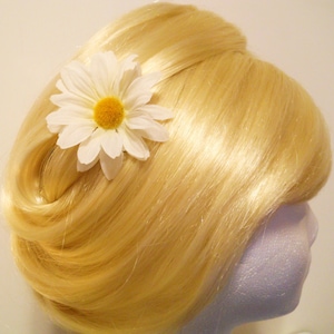 May include: A blonde wig styled with a classic updo, adorned with a large white daisy hair clip. The daisy has a yellow center and white petals. The wig is on a white foam head.