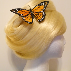 May include: A blonde wig styled with a bouffant and a butterfly hair clip. The butterfly has orange and black wings with white spots. The wig is on a white foam head.