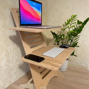 May include: A light-coloured wooden standing desk converter with two tiers. The top tier holds a silver laptop with a colourful screen display. The lower tier features a white wireless keyboard, a black computer mouse, and a black smartphone.