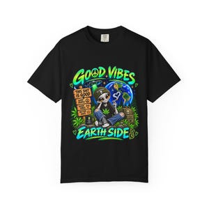 Könnte beinhalten: Ein schwarzes T-Shirt mit einer Grafik eines Außerirdischen, der in einem Wald vor dem Hintergrund der Erde sitzt. Der Text lautet GOOD VIBES, EARTH SIDE, THIS SHIT IS GOOD, GOOD PEOPLE, GOOD VIBES, BETTER DAYS und PEACE LOVE PLANTS.