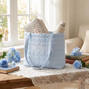 Puede incluir: Un bolso tote a cuadros azul claro y blanco con el nombre "OLIVIA" bordado en la parte delantera. La bolsa contiene una almohada estampada, una botella de agua y otros artículos. La bolsa está sobre una mesa con flores y libros.