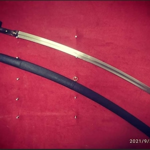 May include: A curved sword with a silver blade and a black handle, alongside a black scabbard. The sword is displayed on a red surface.