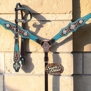 May include: A dark brown leather horse breast collar and headstall set featuring turquoise textured leather overlays, leopard print accents, and clear crystal conchos. A copper-toned centre plate and a Saddle & Stitched brand tag are attached.