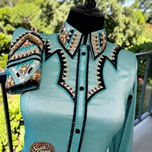 May include: A light blue long-sleeve shirt featuring a pointed collar and front placket adorned with black trim and intricate geometric patterns of brown, clear, and silver crystals. A brown logo patch reads Saddle & Stitched Show Shirts & Blankets.