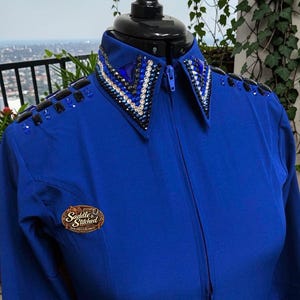 May include: A royal blue long-sleeve shirt featuring a front zipper closure and a pointed collar. The collar and shoulders are embellished with rows of clear, blue, and black crystals. A logo patch reads Saddle & Stitched Show Suits & Blankets.