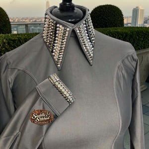 May include: A long-sleeved grey shirt featuring a pointed collar and cuffs embellished with rows of clear and iridescent crystals. The shirt has a smooth, satin-like finish and includes a logo patch reading Saddle & Stitched on the sleeve.