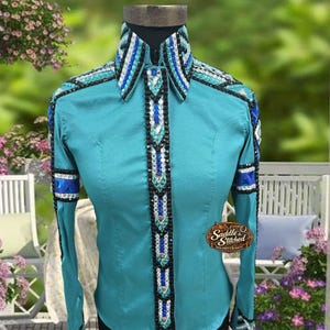 May include: A teal long-sleeve shirt featuring a structured collar and a vertical front placket adorned with intricate rows of blue, white, and black crystals. The sleeves and collar edges are embellished with matching crystal patterns and a logo.