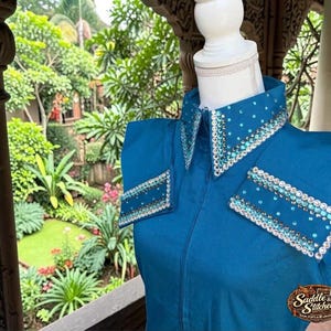 May include: A teal long-sleeve shirt featuring a pointed collar and rectangular shoulder accents adorned with clear and light blue rhinestones. The shirt has a front zipper closure and a small logo patch reading Saddle & Stitched.
