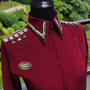 May include: A deep burgundy long-sleeve shirt featuring a zip-up front, a structured collar, and shoulders embellished with rows of clear and dark crystals. The chest is adorned with scattered dark rhinestones and a Saddle & Stitched logo patch.
