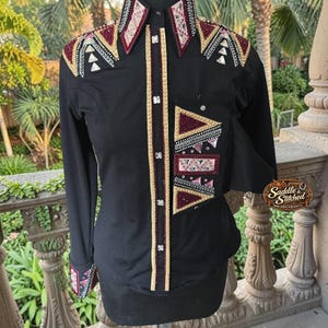 May include: A black long-sleeve shirt featuring a button-down front with gold trim, a pointed collar, and cuffs. The shirt is decorated with geometric patterns in maroon, gold, and silver crystals on the shoulders, collar, and chest pocket.