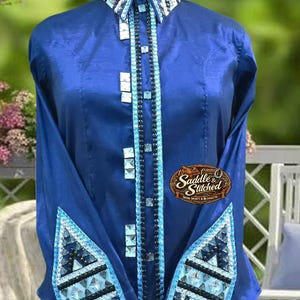 May include: A royal blue long-sleeve shirt featuring a high collar and cuffs adorned with large clear crystals and light blue accents. A central vertical panel displays dark beads and square crystals. A logo patch reads Saddle & Stitched.