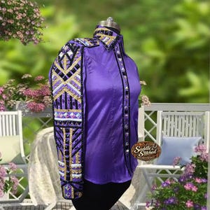 May include: A long-sleeve purple shirt featuring a decorative panel on the sleeve and front placket adorned with black, gold, and white geometric patterns, clear crystals, and black trim. A logo patch reading Saddle & Stitched is attached.
