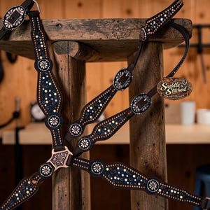 May include: A dark brown leather horse breast collar featuring intricate silver-toned metal conchos, small metallic studs, and a central floral-engraved plate. A brown oval tag with the text Saddle & Stitched is attached to the leather strap.