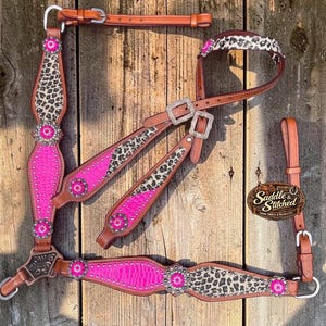 May include: A leather horse headstall and breast collar set featuring bright pink textured panels, leopard print accents, and silver-toned hardware. The set includes decorative floral conchos with pink centers and the text Saddle & Stitched.