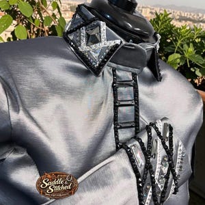 May include: A metallic silver show shirt featuring a high collar adorned with black and clear crystals in a geometric pattern. A vertical row of black rectangular crystals runs down the center, and the sleeve cuff displays a crisscross black bead design.