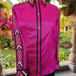 May include: A bright fuchsia long-sleeve button-down shirt featuring a structured collar and cuffs adorned with gold-tone pyramid studs, black trim, and white accents. The front placket is lined with black crystals and gold-tone square studs.