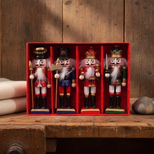 Può includere: Un set di quattro statuette schiaccianoci in legno esposte in una scatola di cartone rossa. Ogni figura presenta un'uniforme dipinta, una barba in pelliccia sintetica bianca e un cappello unico, tra cui copricapi militari e due corone.