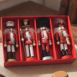 May include: A set of four wooden nutcracker ornaments arranged in a red cardboard box. Each figure wears a red uniform with white accents, black boots, and a white faux fur beard. The figures feature distinct hats, including a tall black bearskin cap.