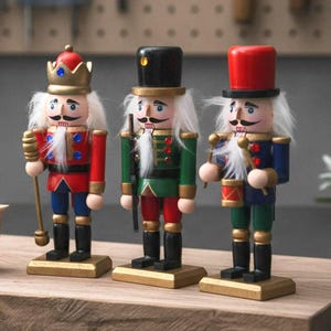 May include: Three wooden nutcracker figurines standing on rectangular bases. The left figure wears a gold crown and red coat, the center figure wears a black top hat and green coat, and the right figure wears a red top hat and blue coat.