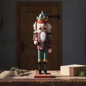 Puede incluir: Una figura de cascanueces de madera con una corona dorada con detalles azules, una chaqueta roja con flecos dorados y pantalones a rayas. La figura tiene una barba de piel sintética blanca, sostiene una lanza dorada y se apoya en una base roja.