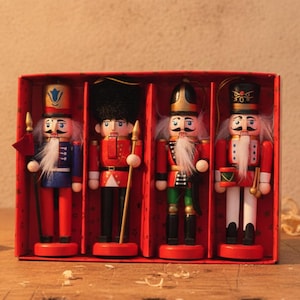 Könnte beinhalten: Ein Set aus vier hölzernen Nussknacker-Anhängern in einer roten Pappschachtel. Jede Figur hat eine bemalte Uniform, einen weißen Kunstfellbart und eine goldene Aufhängeschnur. Die Nussknacker sind in den Farben Rot, Blau, Grün und Weiß gehalten.