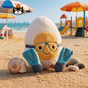 Puede incluir: Un juguete de peluche con forma de huevo con exterior blanco y centro amarillo, que presenta ojos bordados, una pequeña sonrisa, gafas de natación azules y aletas azules en los pies. Una concha en espiral está a la izquierda y guijarros a la derecha.