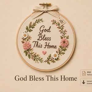 Pode incluir: Um bastidor de bordado circular de madeira contendo tecido creme com o texto God Bless This Home bordado em fio castanho. O texto é rodeado por uma coroa de flores composta por rosas cor-de-rosa, margaridas brancas, folhas verdes e um pequeno coração rosa.