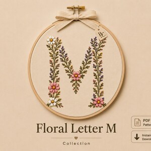 May include: An embroidery hoop containing a cream-colored fabric with the letter M stitched in a floral design. The letter is formed by green stems, leaves, and small pink, white, and purple flowers. A beige ribbon is tied at the top with a small tag.