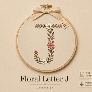 May include: A circular wooden embroidery hoop containing a beige fabric panel with a hand-stitched letter J formed by colourful floral and leaf motifs. A light beige ribbon is tied in a bow at the top, featuring a small tag with the letters BC.