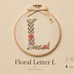 Pode incluir: Um bastidor de bordado circular de madeira contendo tecido cor de creme com uma letra L bordada à mão, formada por flores cor-de-rosa e brancas, folhas verdes e ramos roxos. Uma fita bege está atada no topo com uma pequena etiqueta marcada PC.