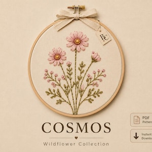 Può includere: Un telaio da ricamo circolare in legno contenente un pannello in tessuto con fiori di cosmo rosa e fogliame verde ricamati a mano. Un fiocco di nastro beige è legato in alto con una piccola etichetta con la scritta BC. Testo COSMOS Wildflower Collection.