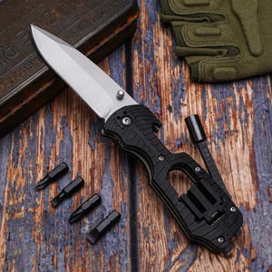 May include: A black and silver folding utility knife with a partially serrated blade, a screwdriver attachment, and several black screwdriver bits. The knife is on a wooden surface.
