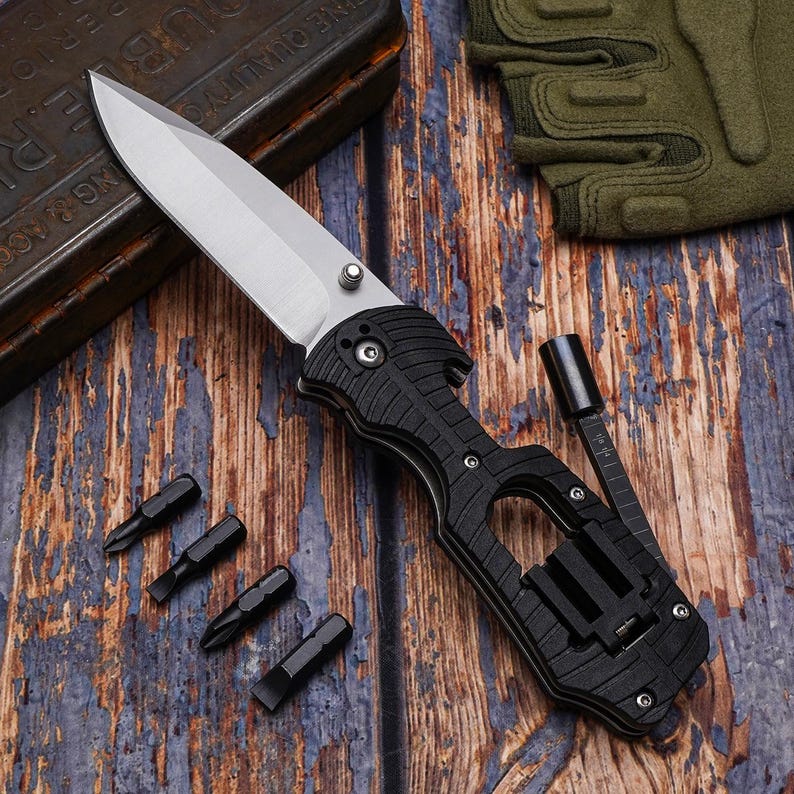 May include: A black and silver folding utility knife with a partially serrated blade, a screwdriver attachment, and several black screwdriver bits. The knife is on a wooden surface.