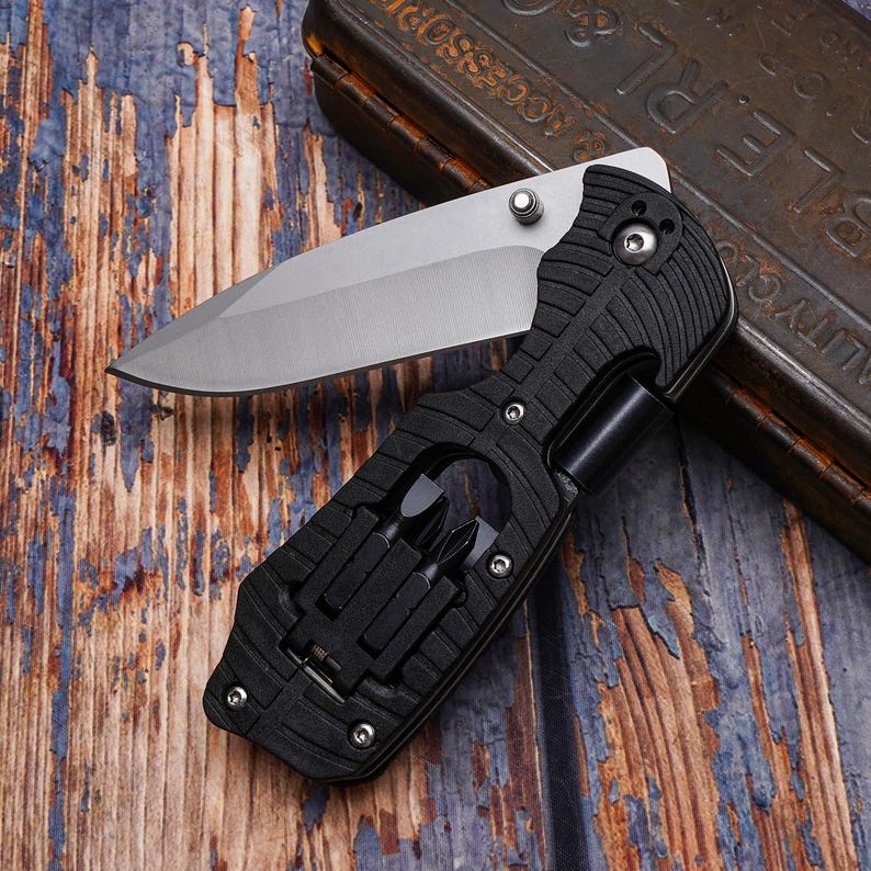 May include: A black multi-tool knife with a partially open silver blade. The handle has a textured grip and includes a set of screwdriver bits. The knife is on a weathered wooden surface.