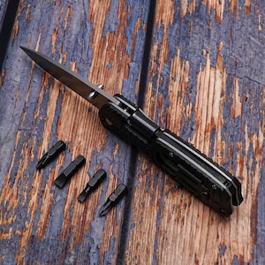 May include: A black multi-tool knife with a silver blade and four black screwdriver bits. The knife is open, revealing the blade. The tools are on a weathered, blue and brown wooden surface.