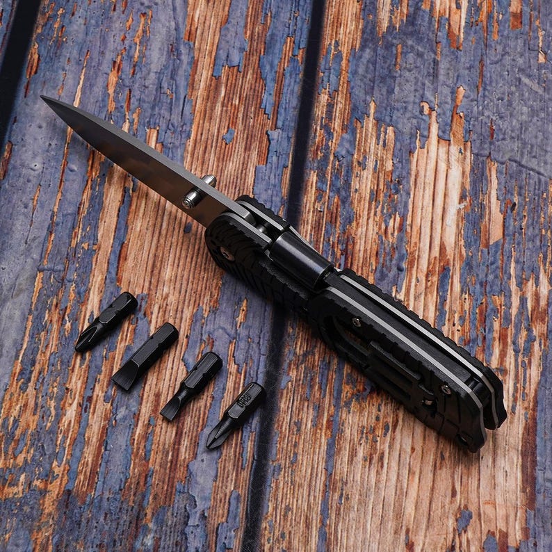May include: A black multi-tool knife with a silver blade and four black screwdriver bits. The knife is open, revealing the blade. The tools are on a weathered, blue and brown wooden surface.