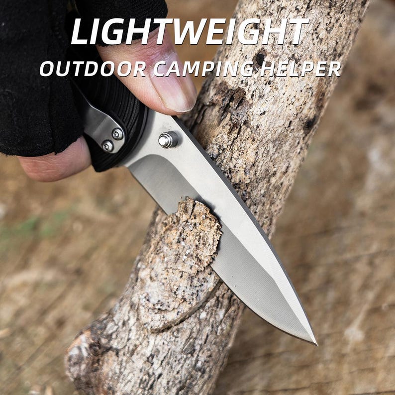 May include: A silver and black folding knife with a sharp blade is cutting into a piece of wood. The text "LIGHTWEIGHT OUTDOOR CAMPING HELPER" is displayed in white letters at the top of the image. The knife is designed for outdoor use.