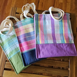 Puede incluir: Tres bolsas de tela rectangulares con asas de tela blanca. Cada bolsa presenta un patrón de cuadros en colores pastel en la sección superior y un panel de tela de color sólido en la parte inferior, incluyendo tonos verde, azul oscuro y morado.