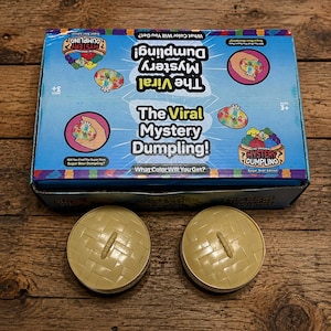 Puede incluir: Una caja de cartón rectangular para The Viral Mystery Dumpling Sugar Bear Edition, con gráficos azules, texto e ilustraciones de dumplings coloridos. Debajo de la caja hay dos recipientes redondos de plástico color canela con textura trenzada.