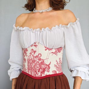 May include: A white off-the-shoulder blouse with ruffled sleeves paired with a cream-colored corset featuring a red toile de jouy print of figures and a donkey. The corset has a red trim at the bottom and is worn over a solid brown skirt.