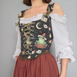 May include: A black corset top featuring a print of frogs wearing hats, teapots, books, and moons. The corset has olive green ribbon lacing down the front, olive green shoulder straps with bows, and is worn over a white off-the-shoulder blouse.