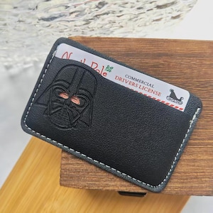 May include: A black leather card holder featuring a debossed Darth Vader helmet design on the front. The card holder has white contrast stitching along the edges and contains a white card labelled North Pole Commercial Drivers License.