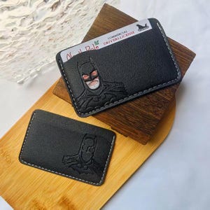 Könnte beinhalten: Zwei schwarze Kartenetuis aus Leder mit geprägtem Batman-Logo. Ein Etui enthält eine weiße Karte mit dem Text North Pole Commercial Drivers License und einem Schlitten-Symbol. Beide Artikel haben weiße Nähte entlang der Kanten.