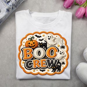 May include: A white t-shirt featuring a scalloped orange border containing the text "BOO CREW" in bold orange and black letters. The design includes embroidered-style graphics of a jack-o'-lantern, two black cats, two ghosts, spiders, and bats.