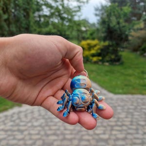 Peut inclure: Une petite figurine d'araignée articulée présentant un dégradé de couleurs bleu métallique et beige. L'araignée possède de grands yeux proéminents, plusieurs pattes segmentées et un anneau métallique fixé sur le dessus du corps pour servir de porte-clés.